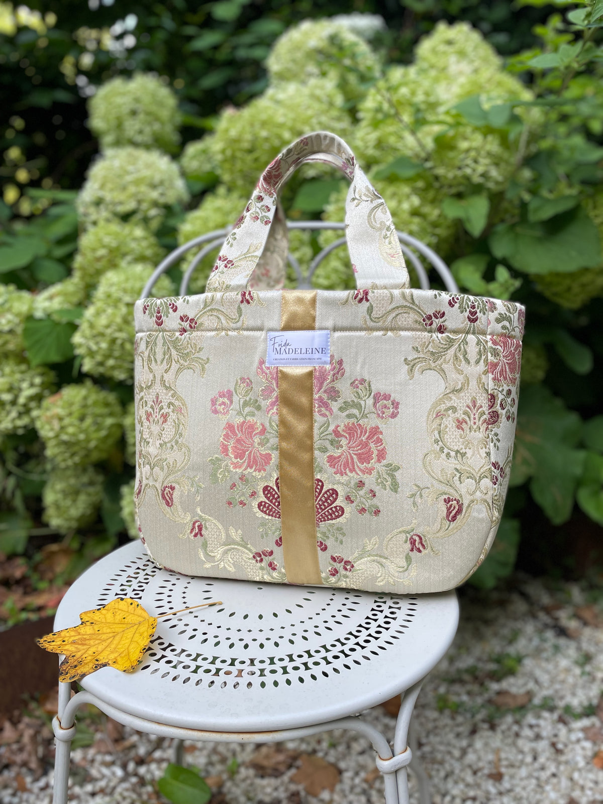 Sac cabas Émile en tissu jacquard au motif baroque doré et rose avec ruban signature Frida Madeleine sur une chaise de jardin